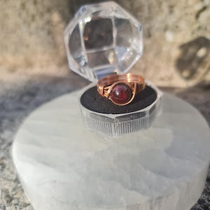 May include: A copper wire ring featuring a round, deep red gemstone, presented in a clear, faceted box. The ring sits on a white, circular stone base. The ring's wirework is detailed, and the gemstone is the central element.