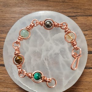 May include: A copper bracelet with various gemstone beads in green, yellow, brown, and dark grey. The beads are linked with copper wire. The bracelet is presented on a white, circular surface. The bracelet's design is intricate.