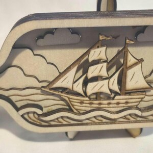 Op de afbeelding: Een gelaagde houten uitsparing in de vorm van een fles met een nautisch tafereel. Het ontwerp toont een zeilschip met witte zeilen op gestileerde golven, met wolken en een vuurtoren op de achtergrond. Het stuk heeft een lichte houten afwerking.