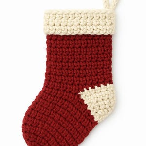 May include: A hand-crocheted Christmas stocking in red, with a cream-coloured cuff and heel. The stocking has a loop for hanging. The textured yarn creates a cosy feel. A festive decoration for the holiday season.