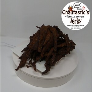 May include: A pile of dark brown beef jerky on a white surface. The jerky is in thin strips and has a slightly spicy look. The text "Chautastic's Small Batch Jerky" is visible in a circle above the jerky.