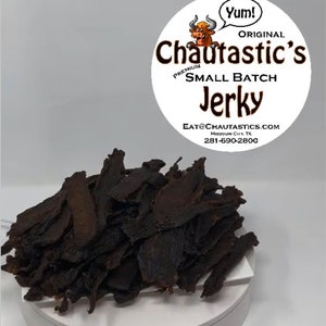 May include: A close-up of a pile of dark brown jerky. The jerky is arranged in a pile on a white surface. A white circle with the text "Yum!" and "Chautastic's Original Premium Small Batch Jerky" is in the background. The text "Eat@Chautastics.com Missouri City, TX 281-690-2800" is also visible in the background.
