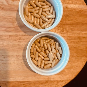 Peut inclure: Deux bols blancs remplis de capsules beiges. Les bols sont sur une surface en bois.