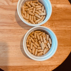 Peut inclure: Deux bols blancs remplis de capsules beiges. Les bols sont sur une surface en bois brun clair.