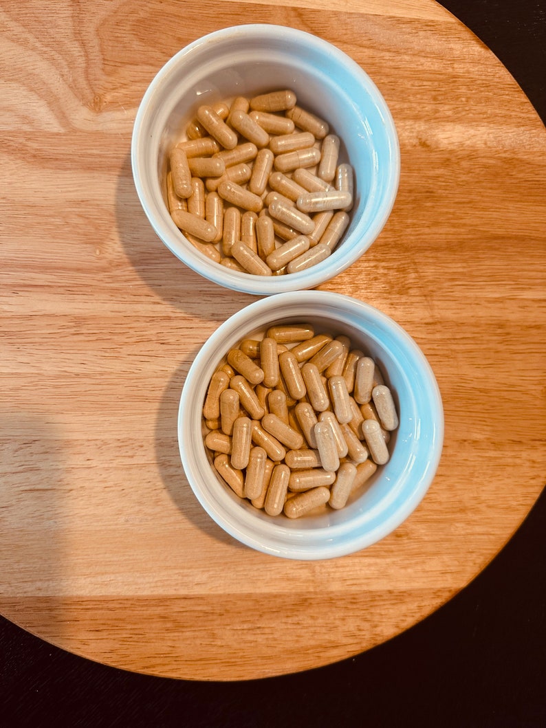 Peut inclure: Deux bols blancs remplis de capsules beiges. Les bols sont sur une surface en bois brun clair.