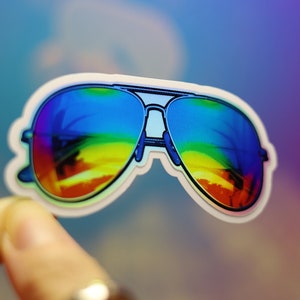 Peut inclure: Un autocollant de lunettes de soleil aviateur avec des verres d&eacute;grad&eacute;s arc-en-ciel. Les lunettes ont une monture argent&eacute;e et un bord blanc. L'autocollant est tenu par un doigt sur un fond flou et color&eacute;.