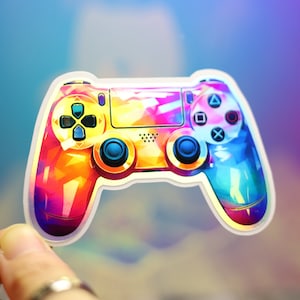 以下が含まれることがあります： カラフルなビデオゲームコントローラーのステッカー。 コントローラーは、オレンジ、黄色、ピンク、青の色合いの鮮やかな幾何学模様が特徴です。 黒いボタンと白い縁があり、背景は青と紫です。