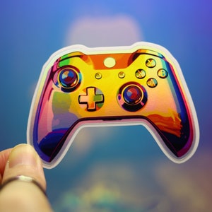 May include: A holographic sticker of a video game controller. The sticker has a rainbow iridescent finish with a yellow center and colorful accents. The controller design includes buttons and a directional pad.