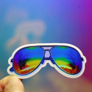 Peut inclure: Autocollant repr&eacute;sentant des lunettes de soleil aviateur avec des verres arc-en-ciel. Les lunettes ont un contour blanc et une monture argent&eacute;e. Le fond est un d&eacute;grad&eacute; de bleu, de violet et de rose.