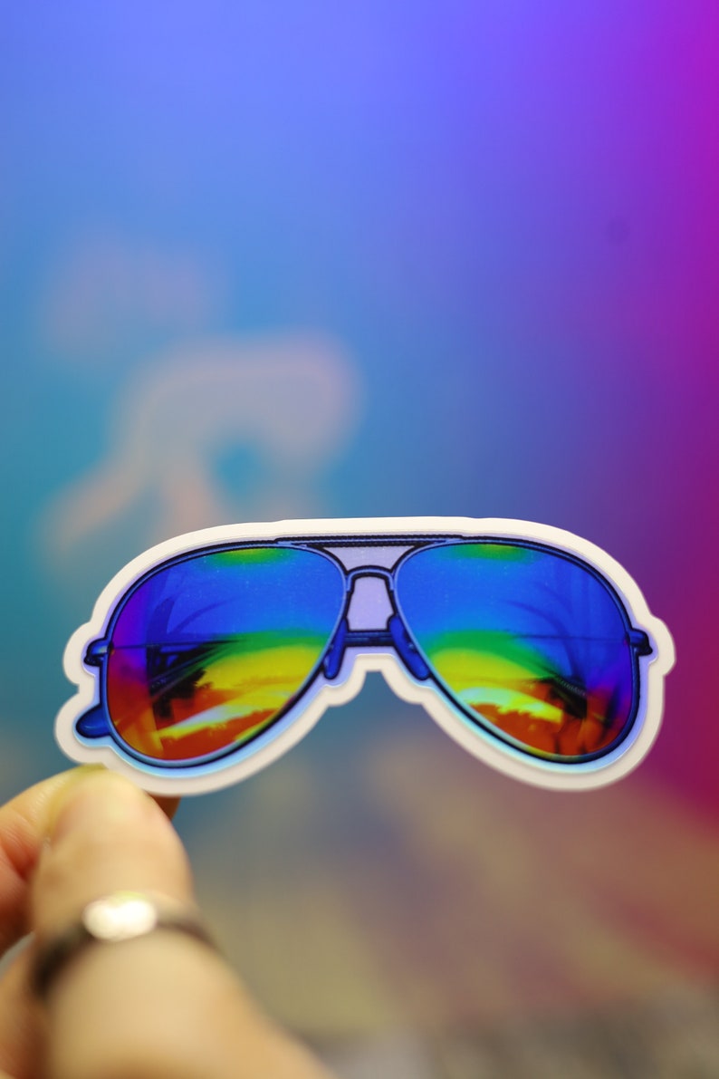 Peut inclure: Autocollant repr&eacute;sentant des lunettes de soleil aviateur avec des verres arc-en-ciel. Les lunettes ont un contour blanc et une monture argent&eacute;e. Le fond est un d&eacute;grad&eacute; de bleu, de violet et de rose.