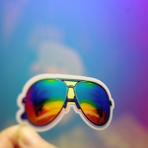 Peut inclure: Un autocollant de lunettes de soleil aviateur avec des verres aux couleurs de l'arc-en-ciel. Les lunettes ont une monture argent&eacute;e et un bord blanc. Les verres refl&egrave;tent une sc&egrave;ne color&eacute;e de plage et de palmiers.