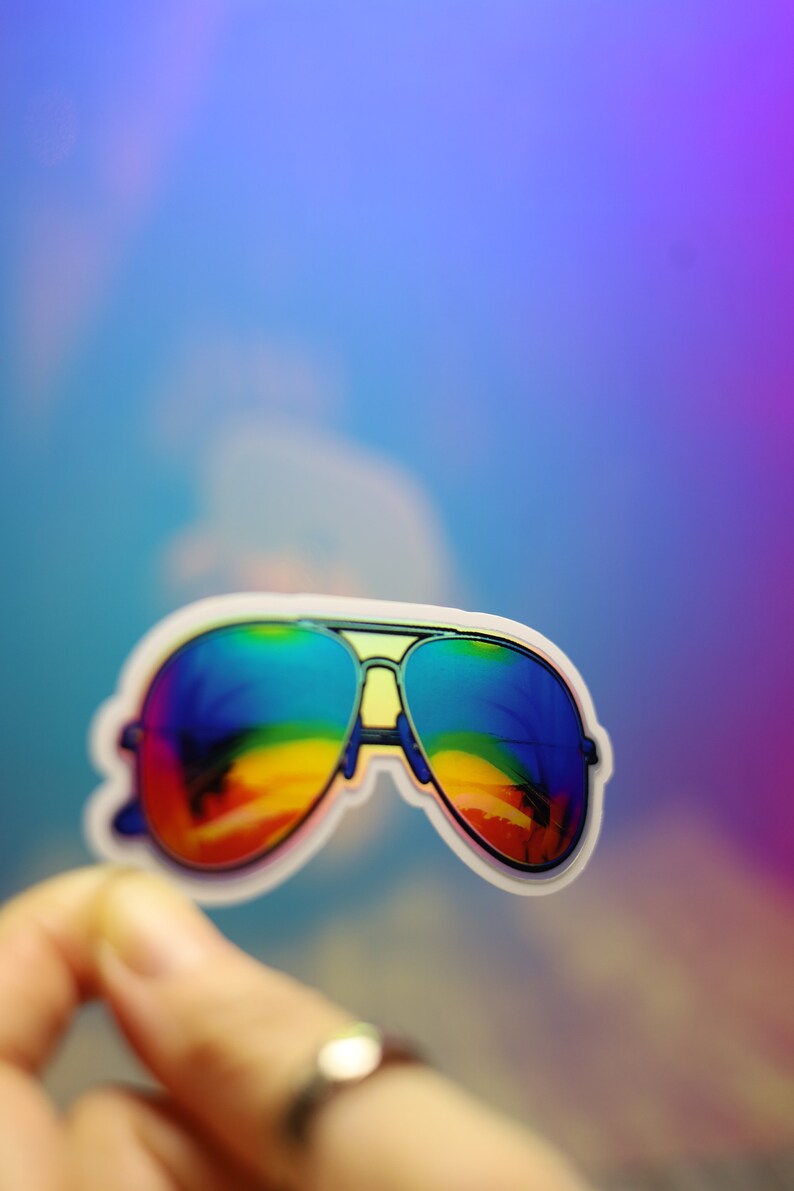 Peut inclure: Un autocollant de lunettes de soleil aviateur avec des verres aux couleurs de l'arc-en-ciel. Les lunettes ont une monture argent&eacute;e et un bord blanc. Les verres refl&egrave;tent une sc&egrave;ne color&eacute;e de plage et de palmiers.