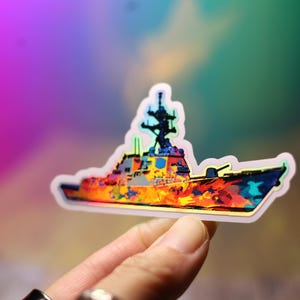 May include: A colorful sticker featuring a stylized warship design. The sticker has a white border and a vibrant, abstract pattern of orange, yellow, blue, and green. The ship's details, including the radar and guns, are visible.