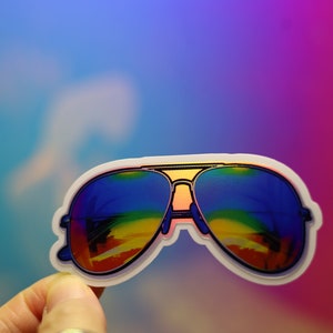 Peut inclure: Un autocollant de lunettes de soleil aviateur avec des verres aux couleurs de l'arc-en-ciel. Les lunettes ont une monture dor&eacute;e et un contour blanc. L'autocollant est pr&eacute;sent&eacute; sur un fond color&eacute; bleu, violet et rose.