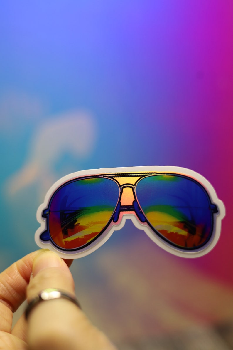 Peut inclure: Un autocollant de lunettes de soleil aviateur avec des verres aux couleurs de l'arc-en-ciel. Les lunettes ont une monture dor&eacute;e et un contour blanc. L'autocollant est pr&eacute;sent&eacute; sur un fond color&eacute; bleu, violet et rose.