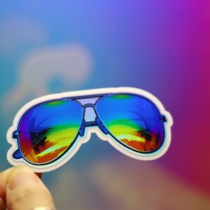 Peut inclure: Autocollant repr&eacute;sentant des lunettes de soleil aviateur avec des verres r&eacute;fl&eacute;chissants arc-en-ciel. Les lunettes ont une monture bleue et un bord blanc. Le fond est un d&eacute;grad&eacute; de bleu, de violet et de rose.