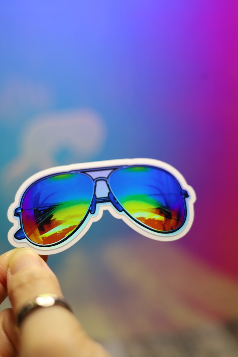 Peut inclure: Autocollant repr&eacute;sentant des lunettes de soleil aviateur avec des verres r&eacute;fl&eacute;chissants arc-en-ciel. Les lunettes ont une monture bleue et un bord blanc. Le fond est un d&eacute;grad&eacute; de bleu, de violet et de rose.