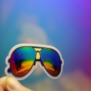 Peut inclure: Un autocollant de lunettes de soleil aviateur avec des verres aux couleurs de l'arc-en-ciel. Les lunettes ont un contour blanc et un pont jaune. Le fond est un d&eacute;grad&eacute; de bleu et de violet.