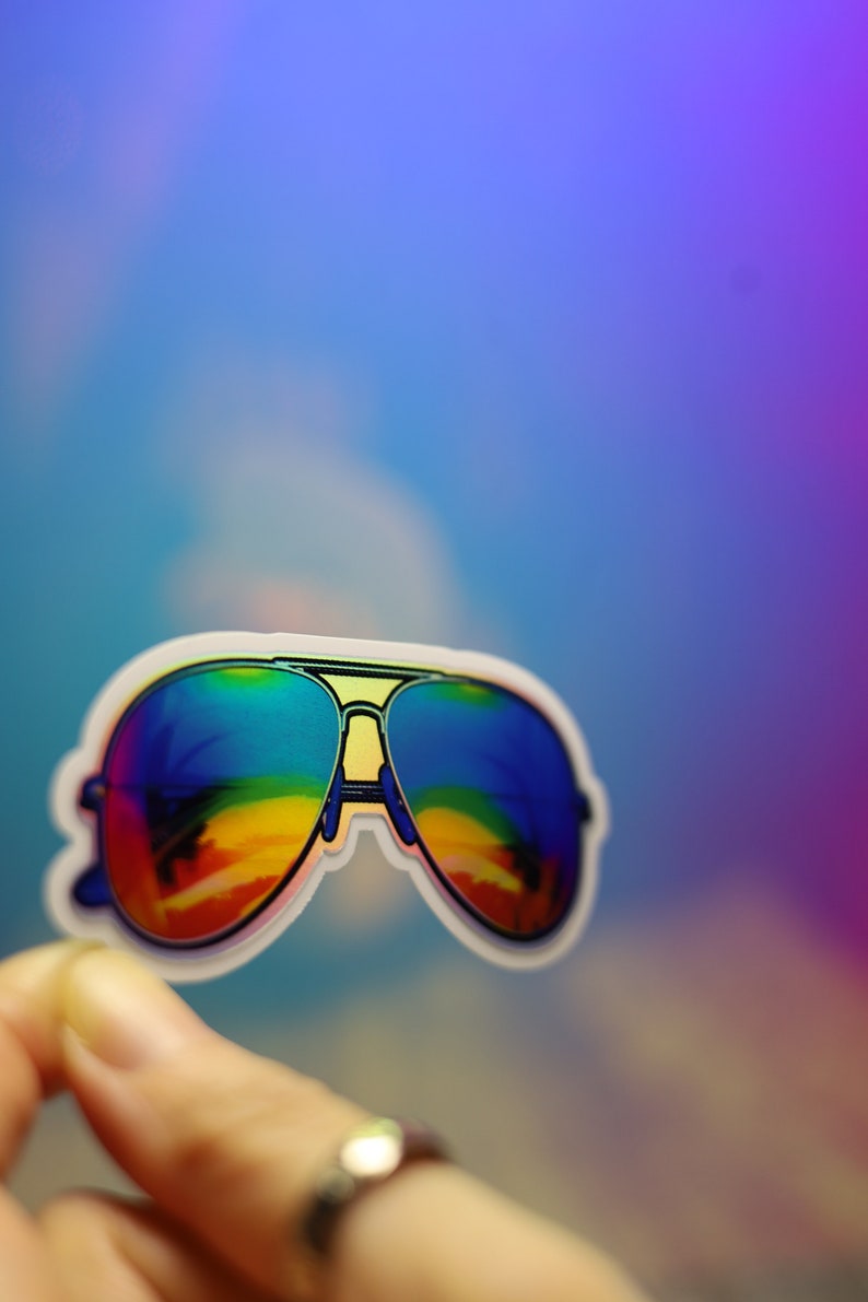 Peut inclure: Un autocollant de lunettes de soleil aviateur avec des verres aux couleurs de l'arc-en-ciel. Les lunettes ont un contour blanc et un pont jaune. Le fond est un d&eacute;grad&eacute; de bleu et de violet.