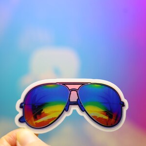 Peut inclure: Un autocollant de lunettes de soleil aviateur avec des verres d&eacute;grad&eacute;s arc-en-ciel. Les lunettes ont une barre frontale rose et un bord blanc. Le fond est un d&eacute;grad&eacute; de bleu, de violet et de rose.