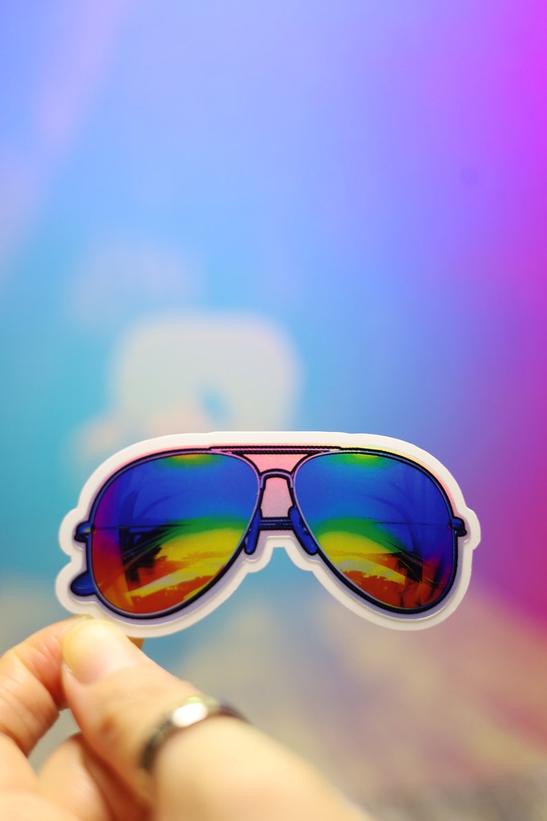 Peut inclure: Un autocollant de lunettes de soleil aviateur avec des verres d&eacute;grad&eacute;s arc-en-ciel. Les lunettes ont une barre frontale rose et un bord blanc. Le fond est un d&eacute;grad&eacute; de bleu, de violet et de rose.