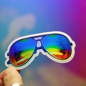Peut inclure: Un autocollant de lunettes de soleil aviateur avec des verres d&eacute;grad&eacute;s arc-en-ciel. Les lunettes ont une monture argent&eacute;e et un bord blanc. L'autocollant est tenu sur un fond color&eacute;.