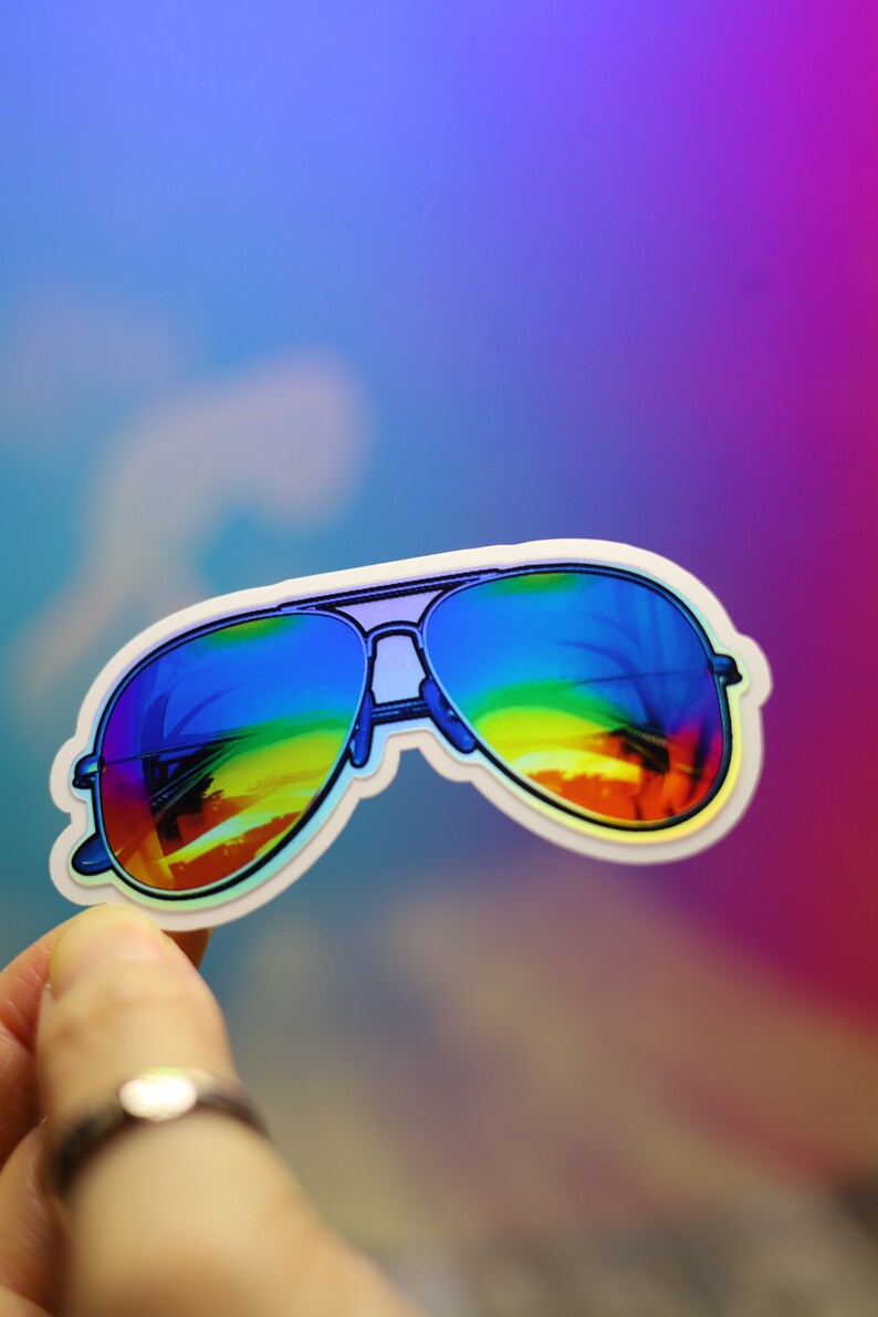 Peut inclure: Un autocollant de lunettes de soleil aviateur avec des verres d&eacute;grad&eacute;s arc-en-ciel. Les lunettes ont une monture argent&eacute;e et un bord blanc. L'autocollant est tenu sur un fond color&eacute;.