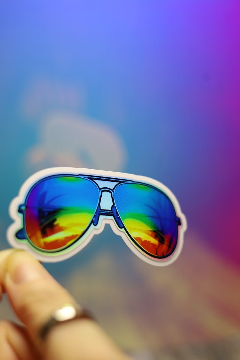 Peut inclure: Un autocollant de lunettes de soleil aviateur avec des verres d&eacute;grad&eacute;s arc-en-ciel. Les lunettes ont une monture argent&eacute;e et un bord blanc. L'autocollant est tenu par un doigt sur un fond flou et color&eacute;.