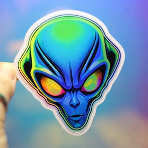 Op de afbeelding: Een sticker met een gestileerde alienkop met een blauw gezicht, groen voorhoofd en iriserende ogen. De alien heeft een strenge uitdrukking en is zwart omlijnd met een witte rand. De achtergrond is een verloop van blauw en paars.