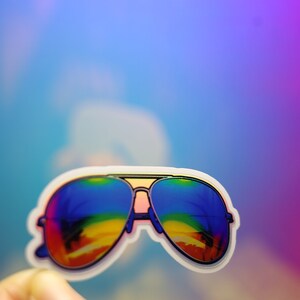 Peut inclure: Un autocollant de lunettes de soleil aviateur avec des verres &agrave; d&eacute;grad&eacute; arc-en-ciel. Les lunettes ont un bord blanc et sont tenues par une main. Le fond est un d&eacute;grad&eacute; de bleu, de violet et de rose.