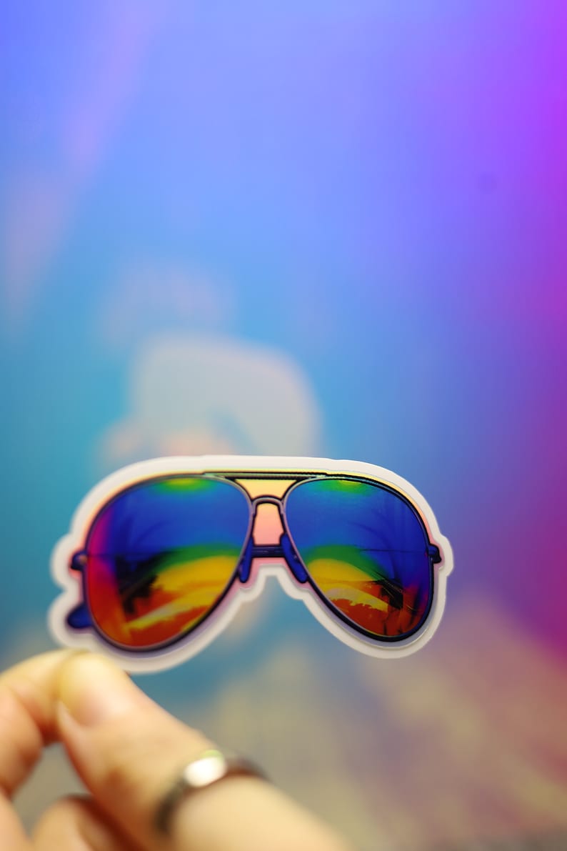 Peut inclure: Un autocollant de lunettes de soleil aviateur avec des verres &agrave; d&eacute;grad&eacute; arc-en-ciel. Les lunettes ont un bord blanc et sont tenues par une main. Le fond est un d&eacute;grad&eacute; de bleu, de violet et de rose.