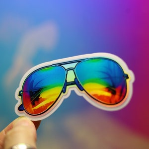Peut inclure: Autocollant repr&eacute;sentant des lunettes de soleil aviateur avec des verres d&eacute;grad&eacute;s arc-en-ciel. Les lunettes ont une monture noire et un bord blanc. Les verres refl&egrave;tent une sc&egrave;ne de coucher de soleil color&eacute;e. L'autocollant est tenu par une main.