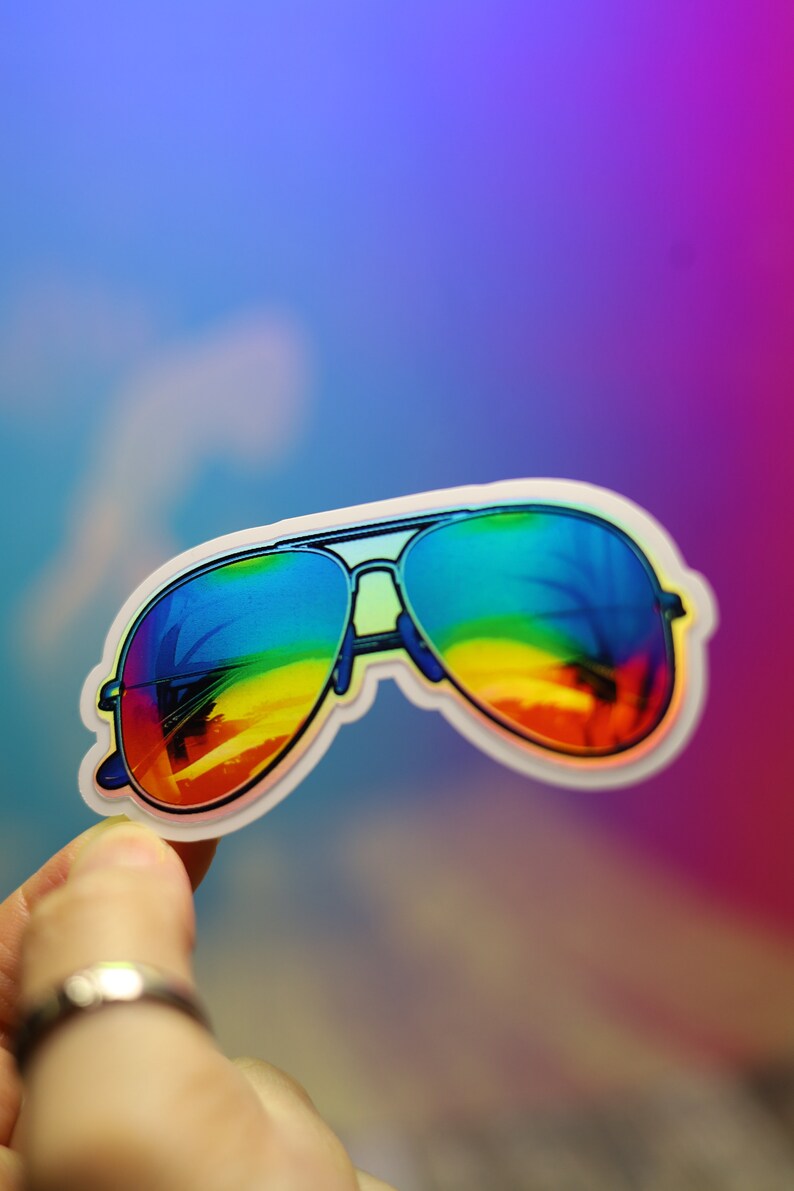 Peut inclure: Autocollant repr&eacute;sentant des lunettes de soleil aviateur avec des verres d&eacute;grad&eacute;s arc-en-ciel. Les lunettes ont une monture noire et un bord blanc. Les verres refl&egrave;tent une sc&egrave;ne de coucher de soleil color&eacute;e. L'autocollant est tenu par une main.