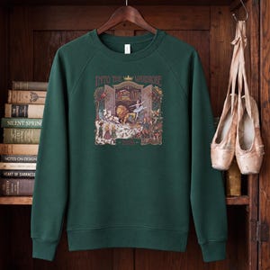 May include: A dark green crewneck sweatshirt featuring a central graphic illustration of a wardrobe, a lion, a ballerina, and children in a snowy forest. The text above the graphic reads INTO THE WARDROBE and the number 2026 is printed below.