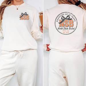 May include: Cream-colored sweatshirt and sweatpants set. The sweatshirt features the letters "NSB" in an orange and black design, with a bow graphic. The back of the sweatshirt has a circular graphic that says "Wrapping Up 10 Years" and "North State Ballet".
