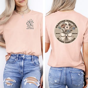 May include: A light pink t-shirt with a gray monogram on the front and a round graphic on the back. The graphic features a bouquet of flowers with a bow and the text "Practice Kindness, Give, Bloom Together, Grow".