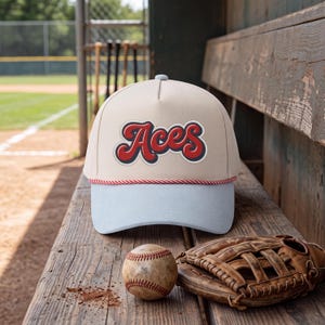 May include: A beige baseball cap with a light blue brim and a red and white twisted cord trim. The front panel features the word Aces in a bold, red, cursive font with a dark blue outline. The cap has a structured five-panel design.