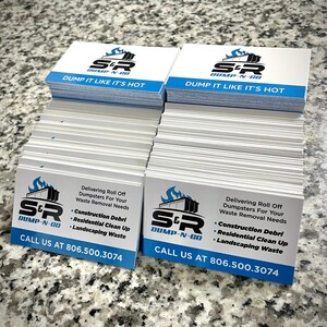 May include: Stacks of white business cards with a blue and black logo for S&R Dump-N-Go. The cards have the text "DUMP IT LIKE IT'S HOT" and list services including construction debris removal and residential clean up. The phone number 806.500.3074 is also displayed.