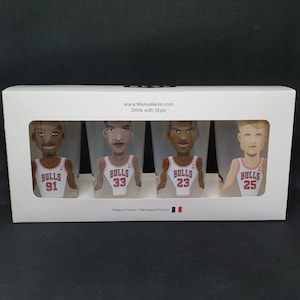 Puede incluir: Conjunto de cuatro vasos de pinta de vidrio transparente con ilustraciones de dibujos animados de jugadores de baloncesto de los Chicago Bulls. Cada vaso presenta a un jugador diferente con un jersey de los Bulls rojo y blanco con su número en la parte delantera. Los números son 91, 33, 23 y 25. Los vasos están en una caja blanca con el texto "www.WeAreReVe.com Drink with Style" y "Made in France/Fabrique en France".