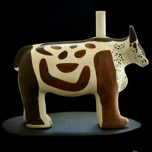 May include: A ceramic sculpture of a cow with a white, brown, and black color scheme. The cow has a cylindrical white piece on its back. The sculpture is on a black surface.