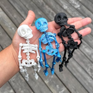 May include: Three small skeleton figurines, one white, one blue, and one black, are held in a person's hand. The skeletons are posed in a standing position with their arms outstretched.