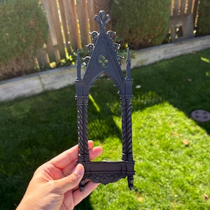 May include: A black gothic decorative frame with detailed carvings, held against a backdrop of green grass and foliage. The frame has a pointed arch design and a fleur-de-lis at the top, reminiscent of a church window.