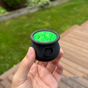 May include: A small, black cauldron with a green, bubbling liquid inside. The cauldron has a rounded shape with a thick rim and a small handle on the side. The green liquid appears to be glowing. The cauldron is held in a person's hand.