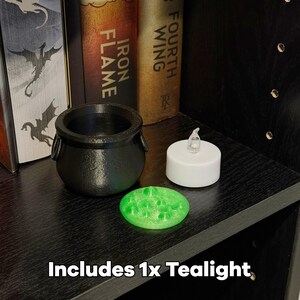 May include: A black cauldron with two handles, a green, textured, circular object, and a white battery-operated tealight. The text "Includes 1x Tealight" is at the bottom. The items are on a black shelf with books in the background.