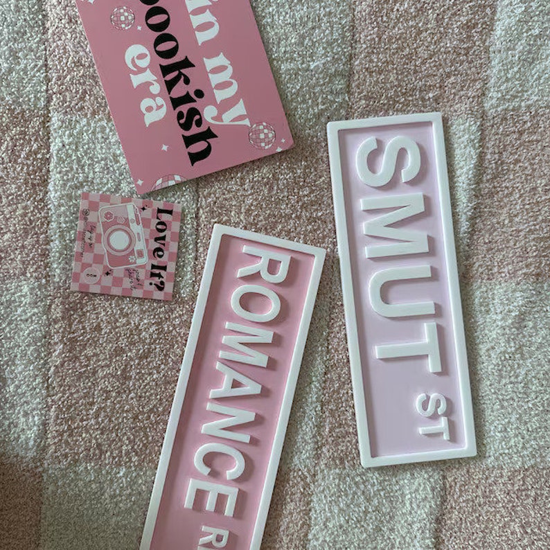 May include: Two pink and white signs with white lettering. The signs read "SMUT ST" and "ROMANCE RD".