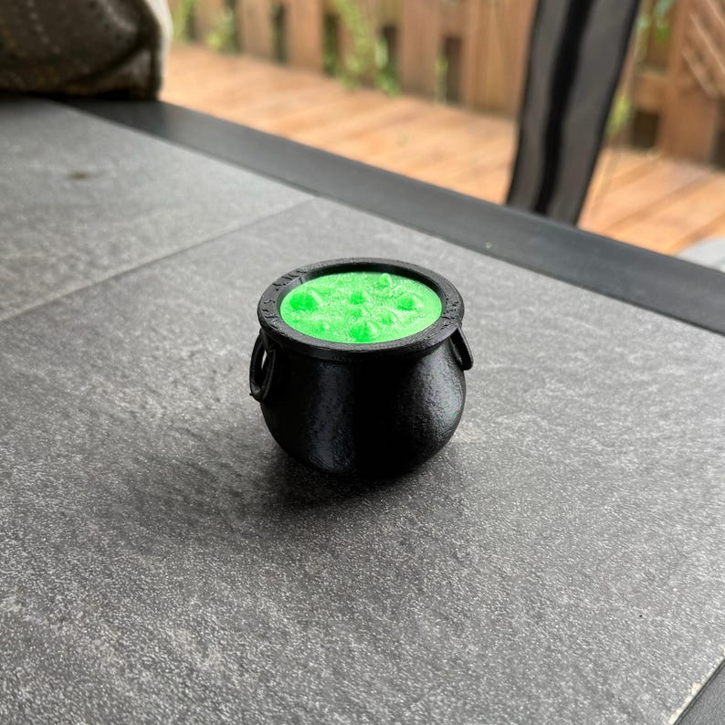 May include: A small, black cauldron filled with bright green liquid. The cauldron has two small handles on the sides and sits on a gray surface. The cauldron is likely used for Halloween or fantasy-themed decorations.