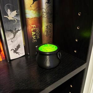May include: A small, black cauldron with a textured surface and two side handles, filled with glowing green liquid. The cauldron sits on a black shelf, next to several books with fantasy-themed covers.