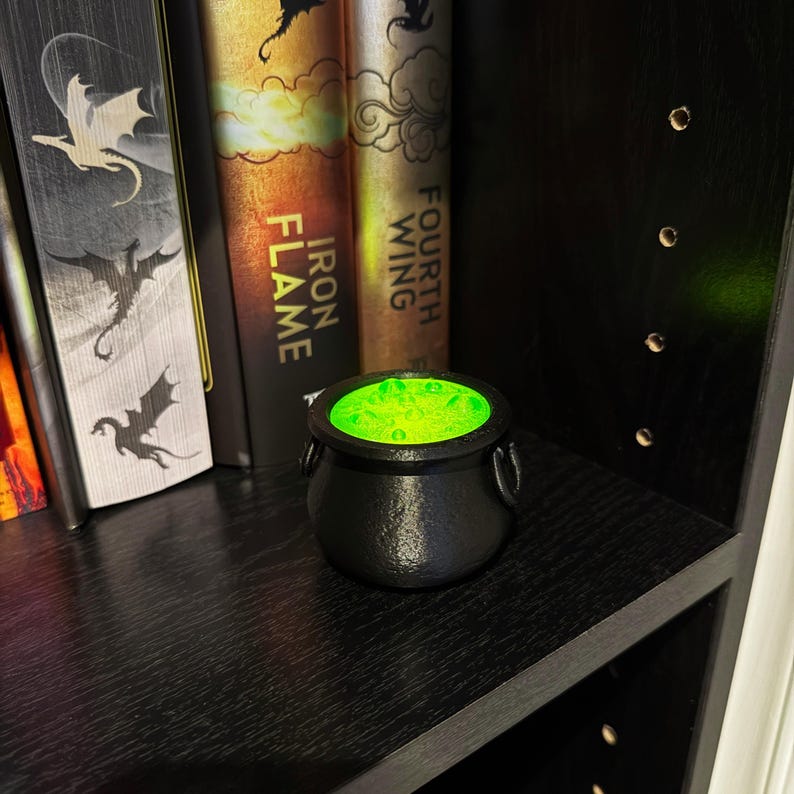 May include: A small, black cauldron with a textured surface and two side handles, filled with glowing green liquid. The cauldron sits on a black shelf, next to several books with fantasy-themed covers.