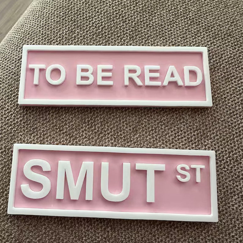 May include: Two pink rectangular signs with white borders and white text. The top sign says "TO BE READ". The bottom sign says "SMUT ST".