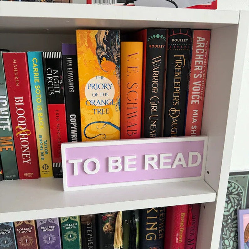 May include: A white bookshelf with a purple sign that reads "TO BE READ". The bookshelf is filled with various books, including "The Priory of the Orange Tree", "Warrior Girl Unear", "Firekeeper's Daughter", and "Archer's Voice".
